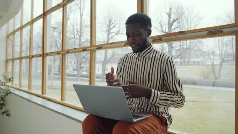 Black-Man-Sitting-on-Window-Sill-and-Video-Calling-on-Laptop